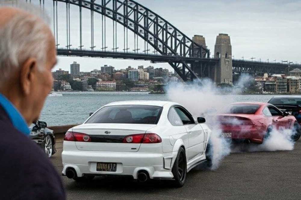 An older man watches three performance cars spinning their wheels in clouds of tyre smoke on a harbourside forecourt, with the Sydney Harbour Bridge behind them.