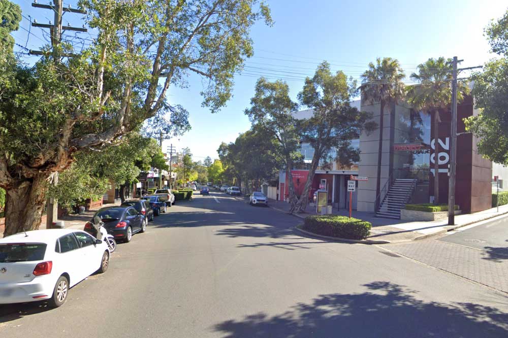 Cars parked nose to tail along the kerb of Blues Point Road, McMahons Point, shaded by tall gum trees, with the office building at 162 Blues Point Road on the corner opposite.
