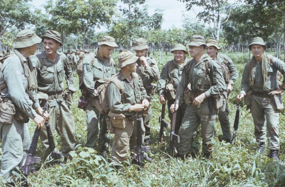 Australian soldiers in jungle greens and bush hats gather in scrub with their rifles during the Vietnam War.