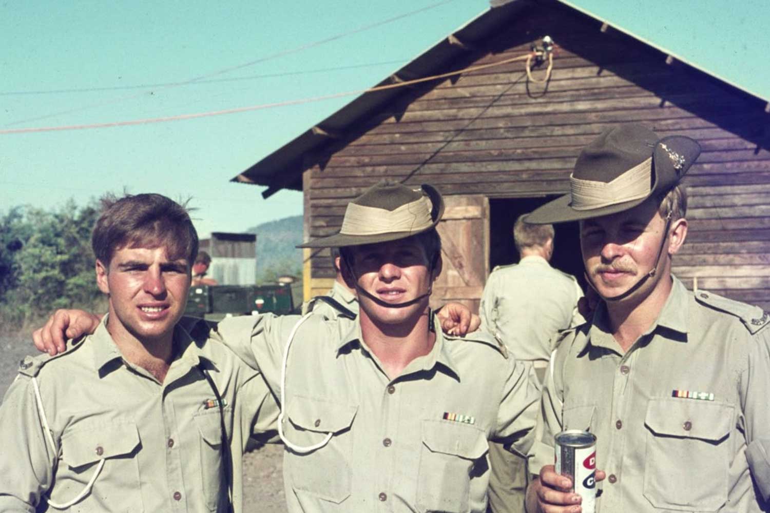 Three young Australian soldiers in slouch hats and jungle greens stand together outside a timber hut during the Vietnam War.