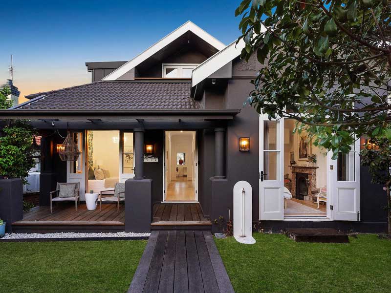 19 Muston St, Mosman The charcoal painted bungalow at 19 Muston St, Mosman, photographed at dusk with open French doors, a timber deck and a lawn path to the front door.