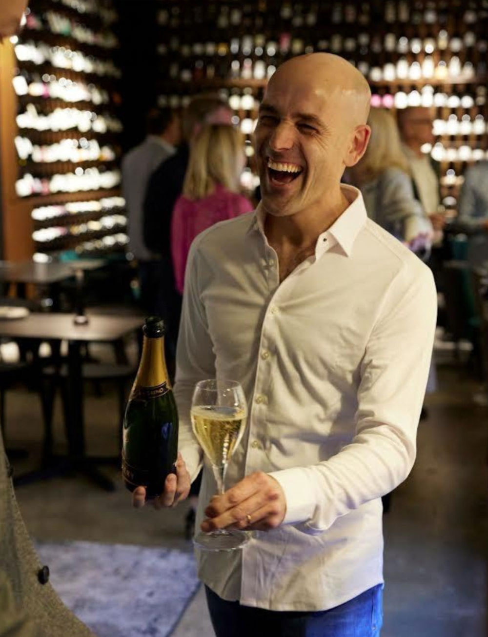 Philippe Gilbert laughing with a bottle of champagne and a glass in a wine bar lined with bottles.