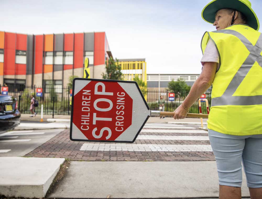A school crossing supervisor holds a Children Crossing stop sign at a pedestrian crossing outside a school.