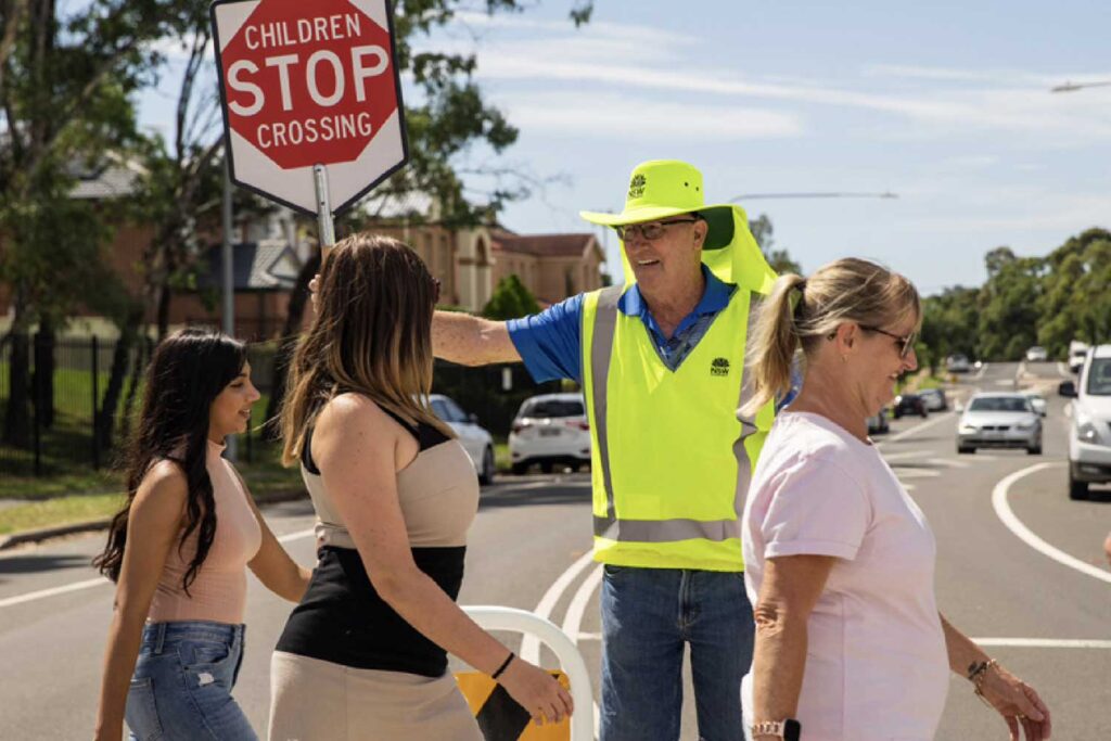 A school crossing supervisor in a high-vis vest and hat guides people across a children's crossing.