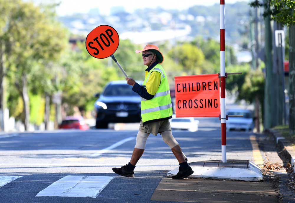 A school crossing supervisor walks across the road holding a stop sign beside a Children Crossing flag.